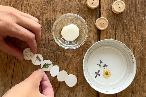 Hands arranging pressed flowers on a wooden surface with small containers and buttons.